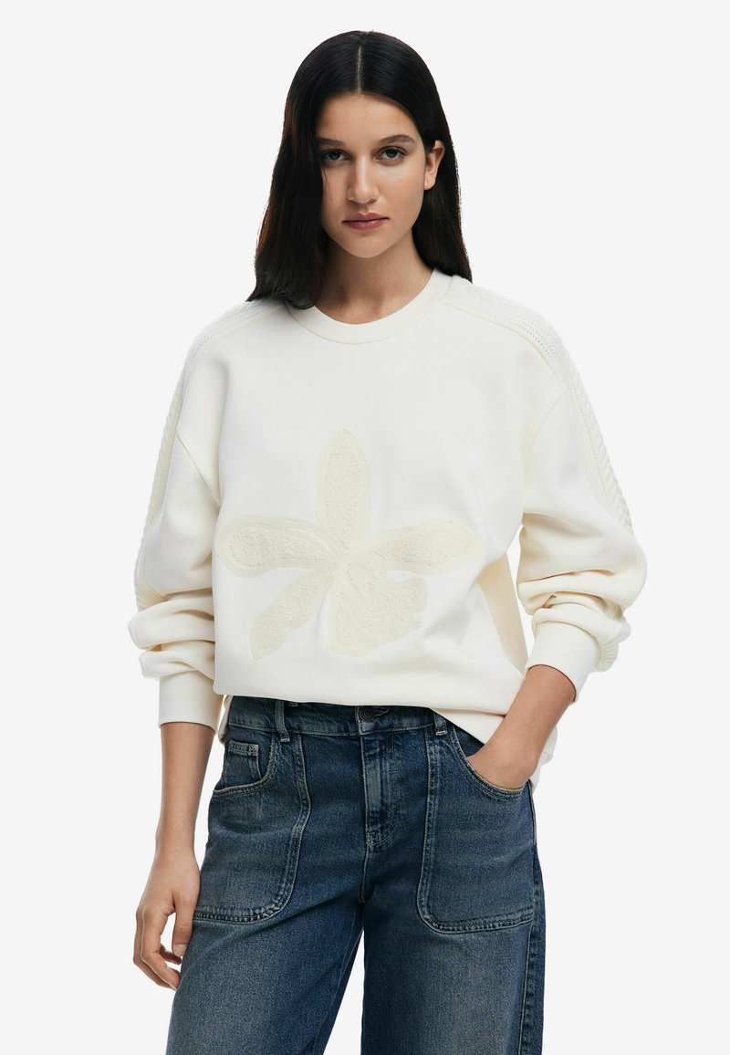 Cream sweatshirt with a textured floral design, featuring ribbed cuffs and a round neckline. Paired with high-waisted, dark blue jeans.