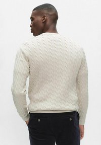 Krämfärgad stickad tröja med flätningsmönster, rund halsringning och ribbade muddar. Texturen är mjuk och något upphöjd. Mörka byxor syns vid midjan.