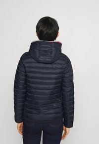 Kvinde med kort mørkt hår iført en mørkeblå quiltet hættejakke og mørke bukser, stående vendt mod en ensartet lys baggrund.