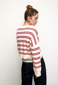 Häkelpullover in Creme mit roten horizontalen Streifen, kurz geschnittenes Design, weite Ärmel und gerippter Saum; kombiniert mit dunklen Jeans.
