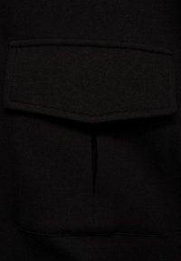 Tissu noir à texture lisse, comprenant une grande poche rectangulaire avec rabat et ouvertures latérales dissimulées.