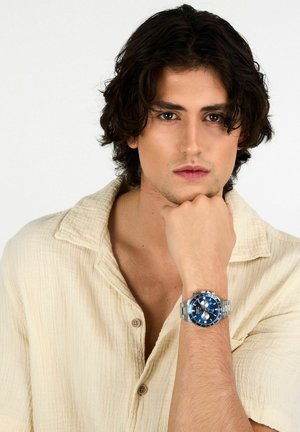 Orologio sportivo con quadrante blu, bracciale in acciaio inossidabile argentato e lancette luminose. Indossato al polso di un modello, con una camicia crema a trama.