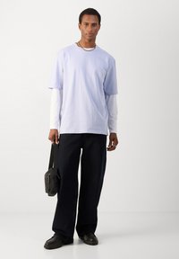 Világos lila rövid ujjú póló, fehér hosszú ujjú ing fölött, sötét bőszárú nadrággal és fekete cipővel párosítva. Fekete postás táska.