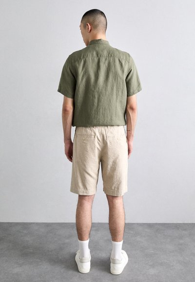 Chemise en lin vert olive, short crème avec taille élastique et poches arrière. Chaussures blanches avec chaussettes blanches.