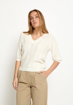 Kvinde med langt blondt hår iført en hvid, struktureret kortærmet sweater og beige bukser med den ene hånd i lommen.