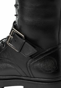 Botte en cuir noir présentant une pointe ronde, un devant lacé, un accent de boucle en argent et un logo embossé sur le côté avec une surface texturée.
