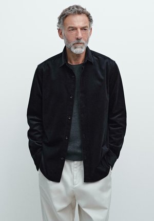 Chemise à boutons en velours côtelé noir avec une surface texturée, associée à un pull gris foncé et un pantalon crème. Mains dans les poches.