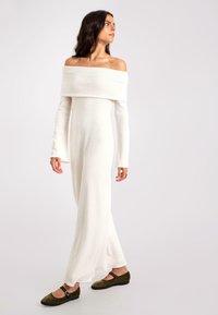 Weißes, schulterfreies langes Kleid mit langen Ärmeln, aus weichem Stoff gefertigt. Fließende Silhouette und taillierter Oberteil, kombiniert mit dunkelgrünen Ballerinas.