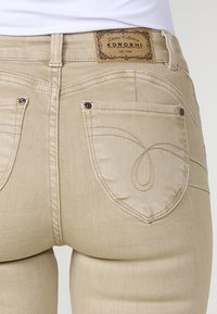 Jeans de mezclilla beige con un diseño distintivo en el bolsillo trasero que presenta costuras curvas y un parche de cuero con el logo en la cintura.