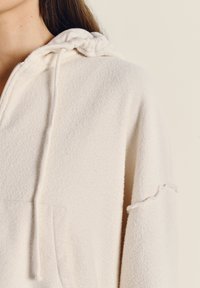 Felpa bianca in pile con superficie testurizzata, dotata di cappuccio con coulisse e tasca frontale. Il design presenta una vestibilità comoda e accenti discreti.