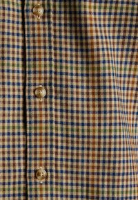  kariertes Hemd in Beige mit blauen, grünen und braunen Quadraten. Glattes Material mit mittlerer Textur. Fünf sichtbare Knöpfe.