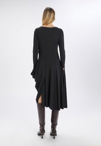 Vestido negro de manga larga con una falda acampanada y bolsillos, que presenta un dobladillo asimétrico y aberturas laterales, combinado con botas marrones hasta la rodilla.