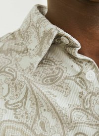 Cămașă bej deschis cu o textură ribbed și un model paisley în nuanțe mai închise de bej. Prezintă un guler ascuțit și închidere cu nasturi.