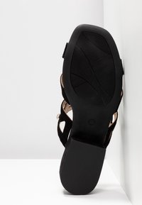 Semelle noire d'une sandale à brides pour femmes avec un talon bloc, présentée debout contre un mur blanc angle.
