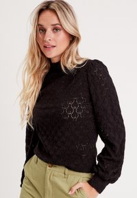 Pull en maille noir avec un motif de losanges texturé, manches longues et col montant ; associé à un pantalon vert olive.