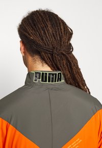 Person med långa bruna dreads som är uppsatta, iklädd en grå och orange Puma-jacka med logotyp på kragen, sedd bakifrån.