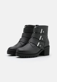 Botas de tobillo negras con un acabado de cuero mate, que presentan tres correas anchas con herrajes plateados y un tacón grueso. Suela de goma texturizada.