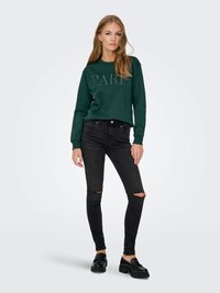 Noor noor wearing a tumeroheline "PARIS" pusak, rebenenud mustade skinny-teksade ja mustade mokassiinide kombinatsioonis, seismas lihtsa heleda tausta ees.
