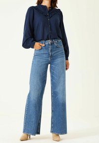 Marinblus med volangkrage, knäppning fram, tillsammans med högmidjade, vida blå denimjeans och beige stilettkängor.