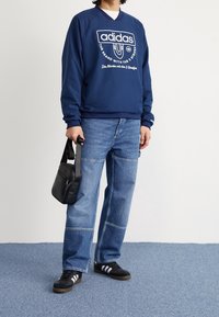 Marinblå sweatshirt med vitt Adidas-logotyp, tillsammans med ljusblå vida jeans, svart axelväska och svarta sneakers med vita ränder.