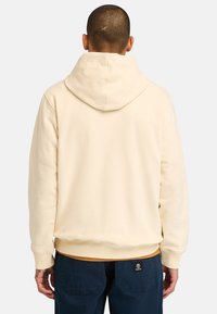 Felpa con cappuccio color crema, dal tessuto morbido, con polsini e orlo a costine, indossata sopra pantaloni blu scuri. Presenta un logo sulla parte posteriore.