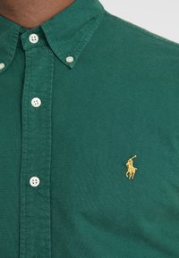 Camisa verde de algodón con botones, que presenta un cuello puntiagudo, botones blancos y un pequeño logo dorado de un jugador de polo en el lado izquierdo.