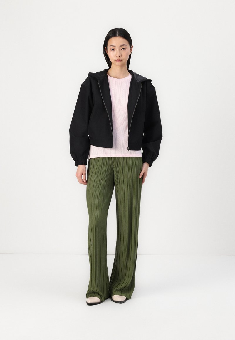 Giacca nera cropped con cappuccio, abbinata a un top a coste rosa chiaro e pantaloni plissettati verdi. Le scarpe sono crema con un accento nero.