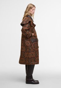 Brauner gemusterter Mantel mit großem Rüschkragen, Seiten Taschen und dunklen Akzenten. Das Material scheint strukturiert zu sein, mit einem floralen Design in warmen Tönen.