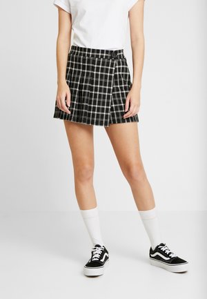 Femme portant une mini-jupe à carreaux noire et blanche, un t-shirt blanc, des chaussettes blanches montantes et des baskets noires basses, debout devant un fond uni.