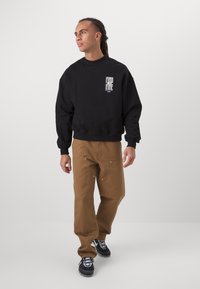 Svart bomullssweatshirt med en hvit logo på brystet, kombinert med brune arbeidsbukser med lommer og detaljer i metall. Svarte joggesko.