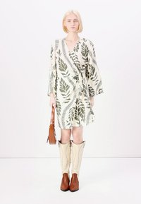 Vestido envolvente en crema con patrón de hojas verdes, mangas de media longitud, cintura ceñida; combinado con botas de cowboy beige y un bolso marrón.