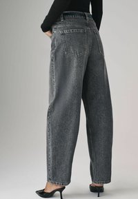 Hoch taillierte, weite Jeans aus verblasstem schwarzem Denim. Verfügt über zwei Gesäßtaschen und eine strukturierte Oberfläche. Kombiniert mit schwarzen High-Heels.