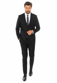 Completo nero con design slim fit, camicia bianca, cravatta nera e scarpe nere lucide. Accentatura a spilla distintiva sul bavero. Aspetto su misura.