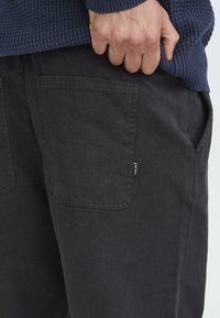 Schwarze Baumwollhosen mit zwei Gesäßtaschen und einem kleinen Markenetikett. Der Stoff hat eine glatte Textur und einen taillierten Schnitt.