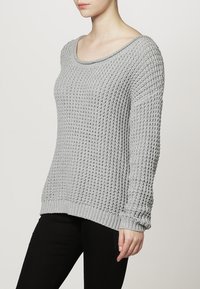 Femme portant un pull ample en maille texturée gris clair à manches longues et un pantalon noir, debout devant un fond blanc.