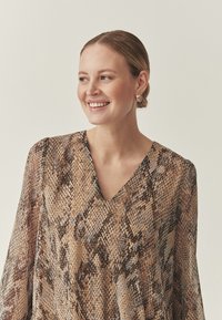 V-Ausschnitt-Bluse mit Schlangenprintmuster in Brauntönen und Beige. Fließende Stoffstruktur mit langen, lockeren Ärmeln. Goldene Creolen.