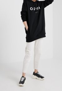 Sweat-shirt à capuche noir avec logo blanc, associé à un pantalon blanc fuselé et des baskets noires avec une semelle épaisse blanche. Look décontracté et sportif.
