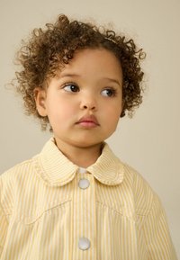 Veste en denim jaune rayée avec un col arrondi et des boutons pression argentés, présentant une texture douce et des coutures visibles.