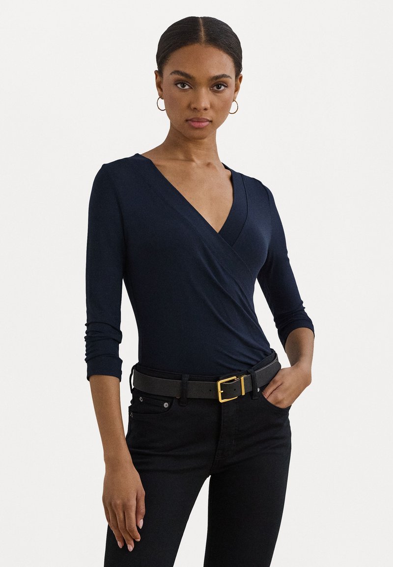 Navy blue long-sleeve wrap top with a deep V-neck, paired with black jeans and a black belt featuring a gold buckle. Smooth fabric texture.