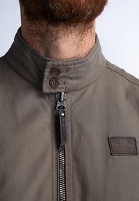 Hnědá bunda s vysokým límcem, vybavená předním zipem, dvěma knoflíkovými uzávěry a koženou nášivkou s detaily značky. Hladká textura.