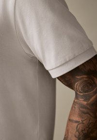 Lys grå polo-shirt med tekstureret stof, foldet ærmekant og synlige sidesømme; båret på en tatoveret arm med intrikate designs.