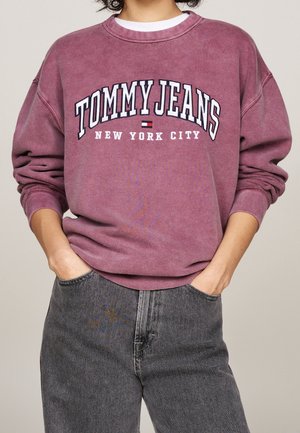 Osoba nosząca wyblakłą fioletową bluzę z napisem i logo „TOMMY JEANS NEW YORK CITY”, zestawioną z szarymi dżinsami z wysokim stanem.