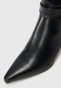 Stivale alla caviglia in pelle nera con punta affusolata, texture liscia e dettaglio di fascia avvolta attorno alla parte superiore. Design minimalista.