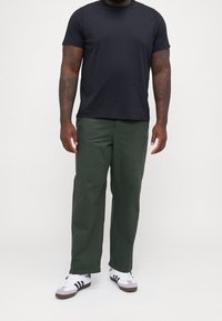 Schwarzes T-Shirt mit kurzen Ärmeln kombiniert mit grünen, weiten Hosen und weißen Sneakers mit schwarzen Streifen. Glatte Stoffqualität mit lockerer Passform.