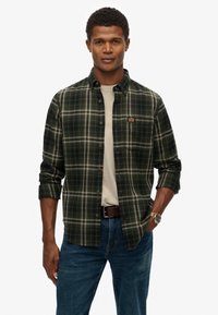 Groene en zwarte geruite blouse met knoopsluiting, kraag en opgerolde mouwen. Draagt over een beige t-shirt, gecombineerd met blauwe jeans.