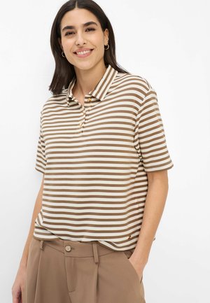 Junge Frau lächelt, trägt ein braun-weiß gestreiftes, kurzärmeliges Poloshirt und hochgeschnittene beige Hose mit den Händen in den Taschen.