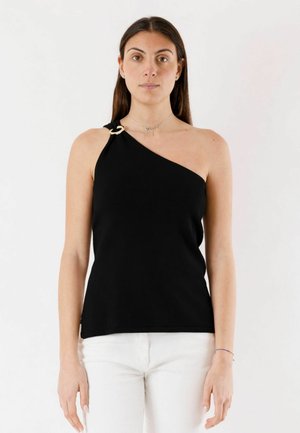 Donna che indossa un top nero monospalla con dettaglio a anello dorato e pantaloni bianchi, in piedi davanti a uno sfondo bianco uniforme.