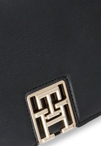 Sort læderhåndtaske med tekstureret overflade, rektangulær form og en fremtrædende guldfarvet metallogo-akkent på forsiden.