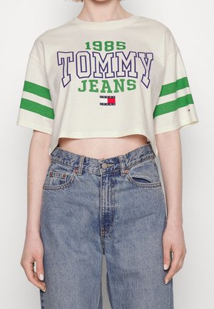 Femme portant un T-shirt blanc court à rayures vertes avec le texte "1985 TOMMY JEANS", associé à un jean denim bleu clair taille haute.