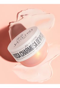 Rosa Dose mit Körpercreme und weißem Etikett mit schwarzem Text. Glatte Creme neben der Dose auf einem weichen rosa Hintergrund verschüttet.